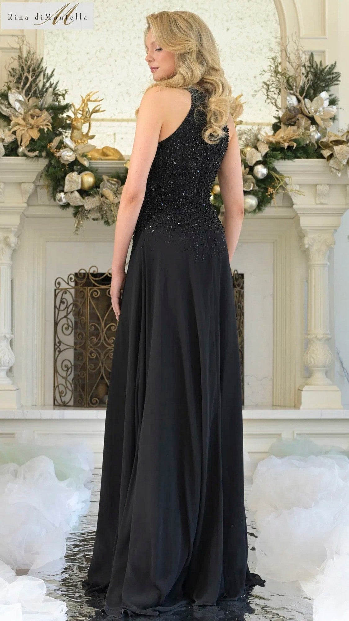 A woman with long blond hair, wearing the Rina Di Montella RD3304 long formal dress, stands with her back to the camera in front of a decorated white fireplace.